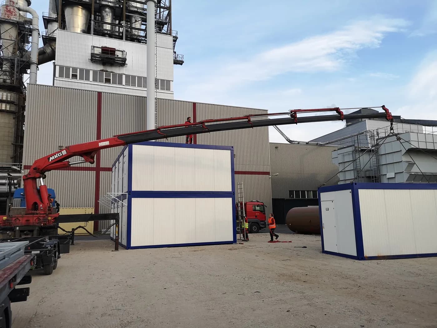 Containeraufbau bei Industrieanlage – Vermietung und Verkauf von Raumcontainern. Montage von modularen Containern mit einem Kran vor einer Industrieanlage – ideal zur Miete oder zum Kauf als Büro- oder Aufenthaltscontainer.