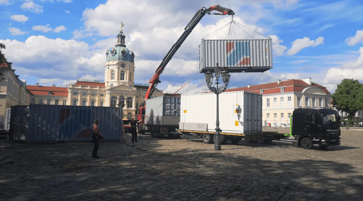 Ein Kran hebt einen Lagercontainer der Mobil-Raum GmbH auf dem Schlossplatz vor dem Schloss Charlottenburg in Berlin, umgeben von weiteren Lagercontainern und Arbeitern.